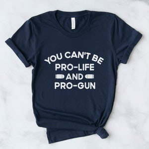 Peut inclure: T-shirt bleu marine avec l'inscription blanche : "YOU CAN'T BE PRO-LIFE AND PRO-GUN", avec des graphiques de balles. Le texte est centr&eacute;. Le t-shirt est pli&eacute; en bas.