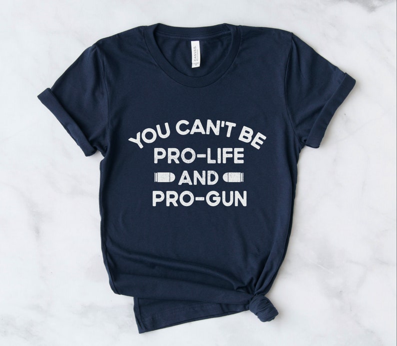 Peut inclure: T-shirt bleu marine avec l'inscription blanche : "YOU CAN'T BE PRO-LIFE AND PRO-GUN", avec des graphiques de balles. Le texte est centr&eacute;. Le t-shirt est pli&eacute; en bas.