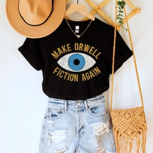 A black unisex crew neck Bella Canvas brand t-shirt that says "MAKE ORWELL FICTION AGAIN" in orange text with a black outline, around an image of a blue eye. The graphic has a distressed vintaged look.