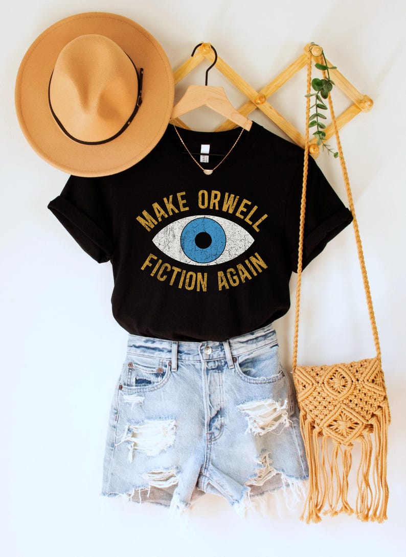 A black unisex crew neck Bella Canvas brand t-shirt that says "MAKE ORWELL FICTION AGAIN" in orange text with a black outline, around an image of a blue eye. The graphic has a distressed vintaged look.
