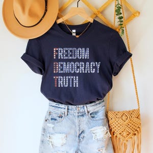 A navy blue unisex crew neck short sleeve Bella+Canvas t-shirt that says "FREEDOM DEMOCRACY TRUTH" on the chest in three lines in blue block text with white stars inside the letters. The first letter of each word in in red with white stars, subtly spelling "FDT" down the shirt