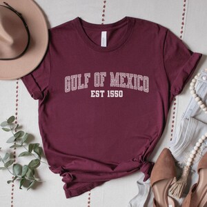A maroon unisex crew neck short sleeve Bella+Canvas brand t-shirt that says "GULF OF MEXICO" on the chest in white outline college style font with white stripes inside the letters. The text is curved over smaller solid white letters in the same font that say "EST 1550". The graphic has a distressed/vintage look