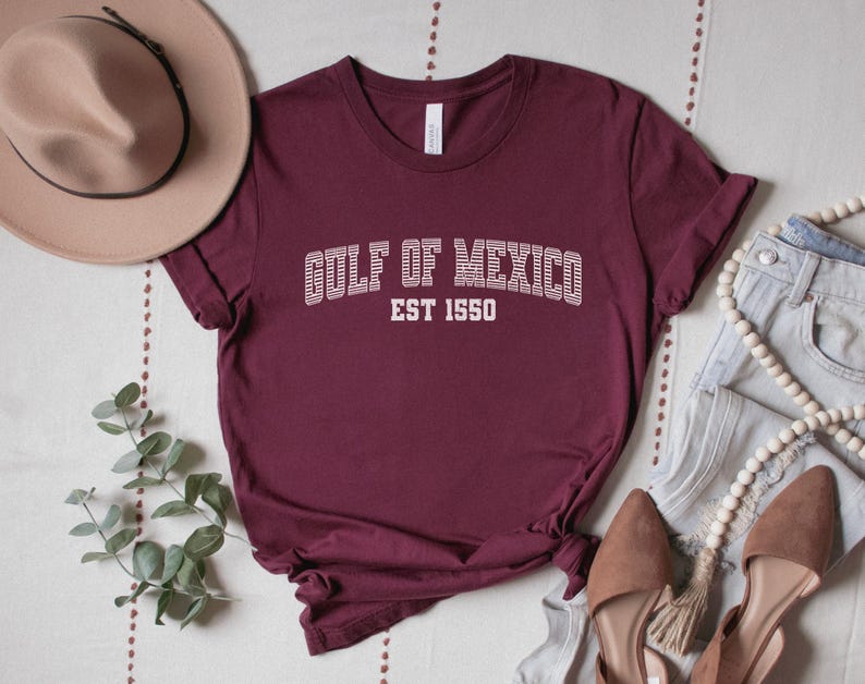 A maroon unisex crew neck short sleeve Bella+Canvas brand t-shirt that says "GULF OF MEXICO" on the chest in white outline college style font with white stripes inside the letters. The text is curved over smaller solid white letters in the same font that say "EST 1550". The graphic has a distressed/vintage look
