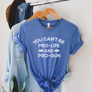 Peut inclure: Un t-shirt bleu avec l'inscription blanche "YOU CAN'T BE PRO-LIFE AND PRO-GUN". Le t-shirt est suspendu &agrave; un cintre en bois. Une chemise en jean et un chapeau marron sont &eacute;galement visibles.