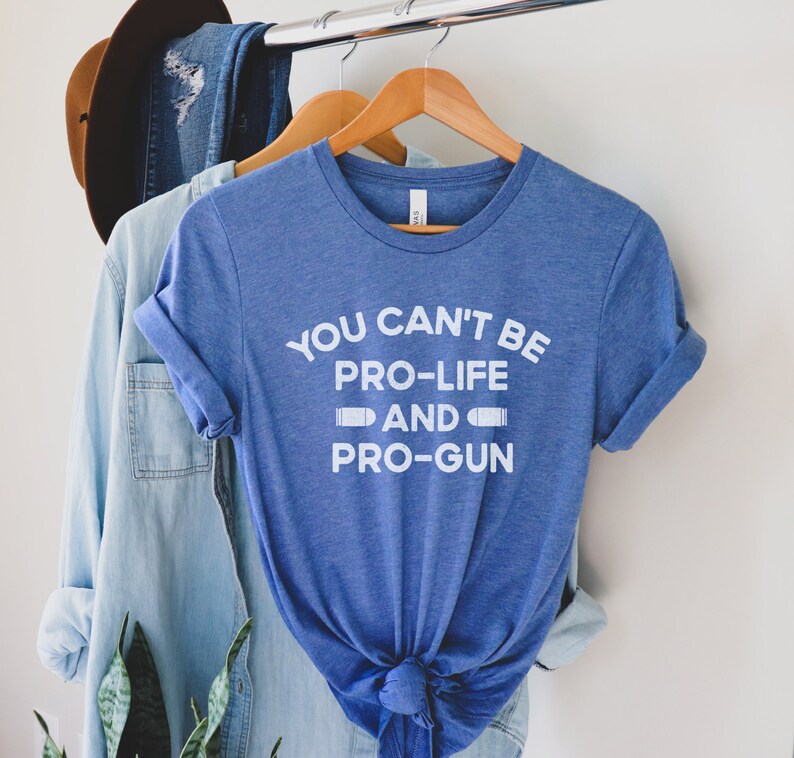 Peut inclure: Un t-shirt bleu avec l'inscription blanche "YOU CAN'T BE PRO-LIFE AND PRO-GUN". Le t-shirt est suspendu &agrave; un cintre en bois. Une chemise en jean et un chapeau marron sont &eacute;galement visibles.