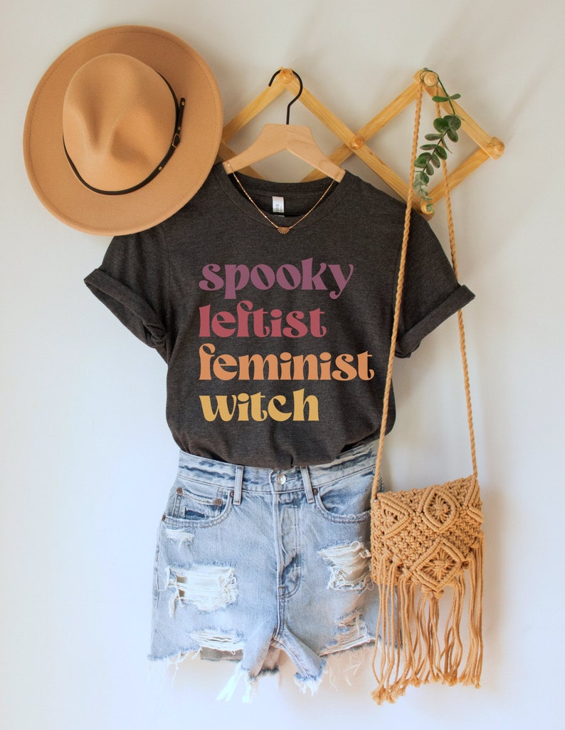 May include: A dark gray t-shirt with the text "spooky leftist feminist witch" in colorful retro font. The shirt is worn with denim shorts and a brown crocheted bag.
