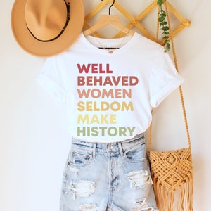 May include: White t-shirt with the text "WELL BEHAVED WOMEN SELDOM MAKE HISTORY" printed in a gradient of red, orange, yellow, and green.