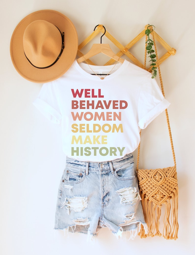 May include: White t-shirt with the text "WELL BEHAVED WOMEN SELDOM MAKE HISTORY" printed in a gradient of red, orange, yellow, and green.