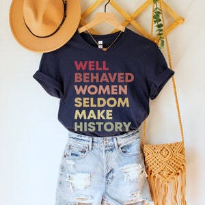 May include: A navy blue t-shirt with the text "WELL BEHAVED WOMEN SELDOM MAKE HISTORY" in a gradient of red, orange, yellow, and gold.