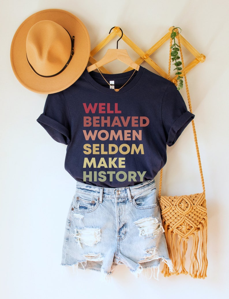 May include: A navy blue t-shirt with the text "WELL BEHAVED WOMEN SELDOM MAKE HISTORY" in a gradient of red, orange, yellow, and gold.