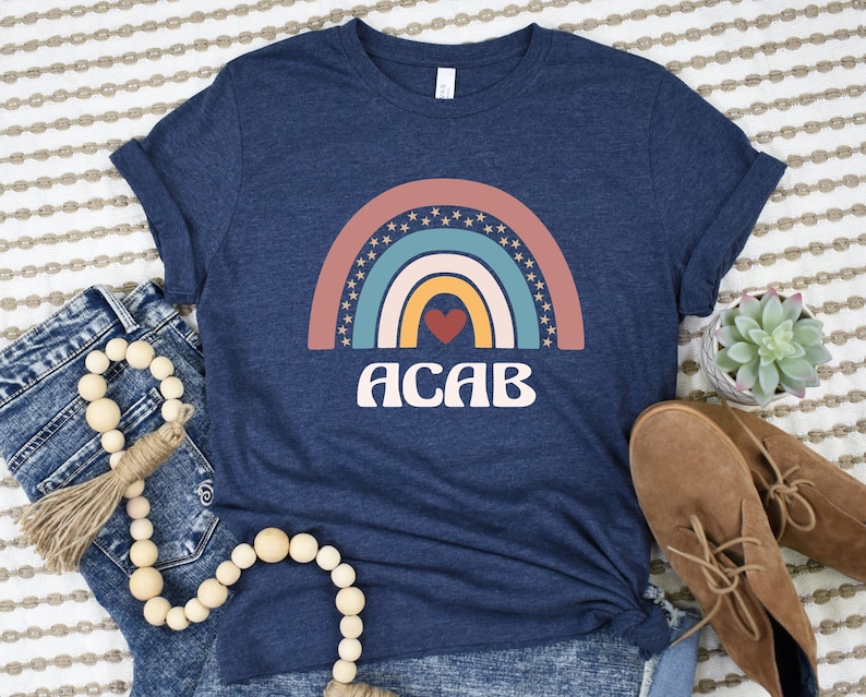 May include: A navy blue t-shirt with a rainbow graphic and the text "ACAB" in a retro font. The rainbow has a red heart in the center.