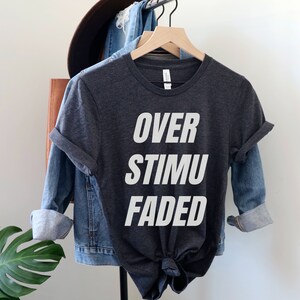 May include: Dark gray t-shirt with the words "OVER STIMU FADED" in large white block letters. The shirt is hanging on a wooden hanger, with a denim jacket and a brown hat in the background.