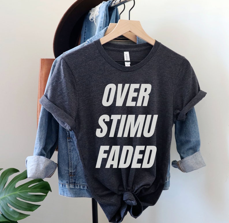 May include: Dark gray t-shirt with the words "OVER STIMU FADED" in large white block letters. The shirt is hanging on a wooden hanger, with a denim jacket and a brown hat in the background.