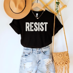 A black unisex crew neck short sleeve Bella+Canvas t-shirt that says "RESIST" in distressed white tall block text