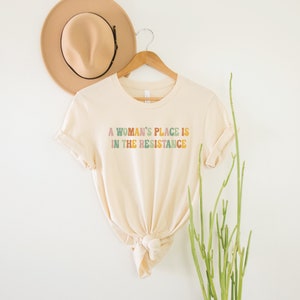 May include: A beige t-shirt with the text "A woman's place is in the resistance" printed in colourful letters.