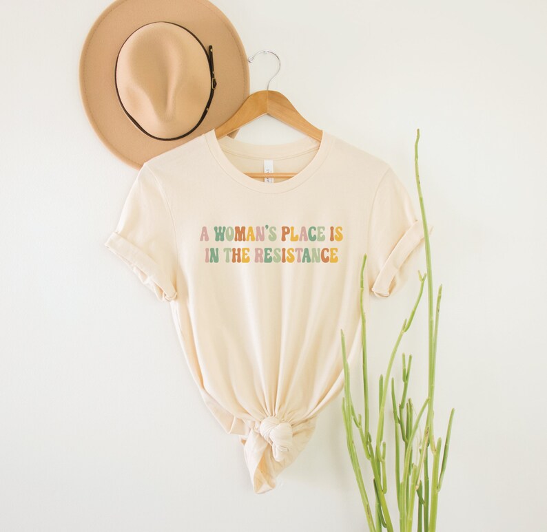 May include: A beige t-shirt with the text "A woman's place is in the resistance" printed in colourful letters.