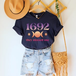 Puede incluir: Una camiseta azul marino con un gr&aacute;fico rosa y naranja del n&uacute;mero 1692 y tres lunas crecientes con estrellas. El texto "they missed one" est&aacute; impreso debajo del gr&aacute;fico.