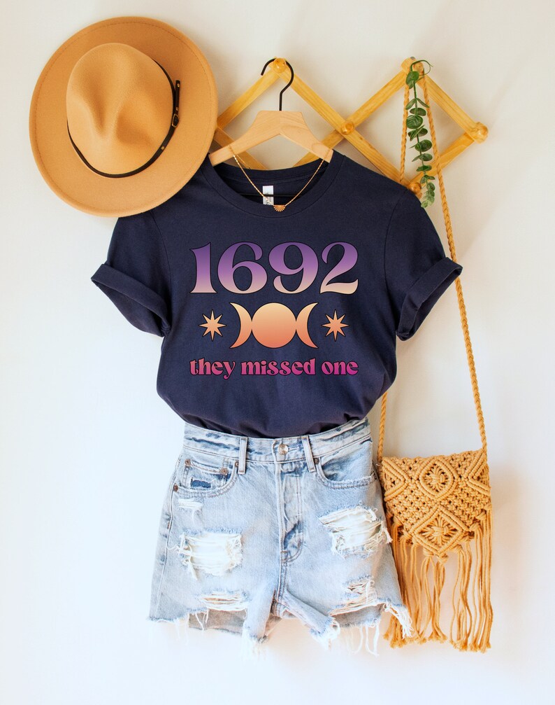 Puede incluir: Una camiseta azul marino con un gr&aacute;fico rosa y naranja del n&uacute;mero 1692 y tres lunas crecientes con estrellas. El texto "they missed one" est&aacute; impreso debajo del gr&aacute;fico.