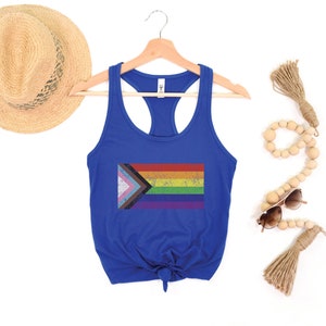 May include: A royal blue tank top with a tie knot at the bottom. The tank top features a distressed, rainbow pride flag with a pink, white, and blue triangle in the top left corner.