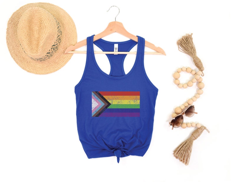 May include: A royal blue tank top with a tie knot at the bottom. The tank top features a distressed, rainbow pride flag with a pink, white, and blue triangle in the top left corner.