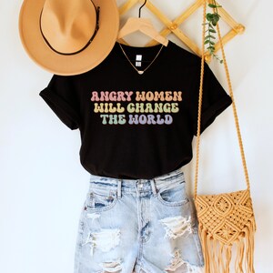 Może przedstawiać: Czarny T-shirt z napisem "Angry Women Will Change The World" w kolorach tęczy. Koszulka wisi na drewnianym wieszaku z brązowym kapeluszem i słomianą torbą.