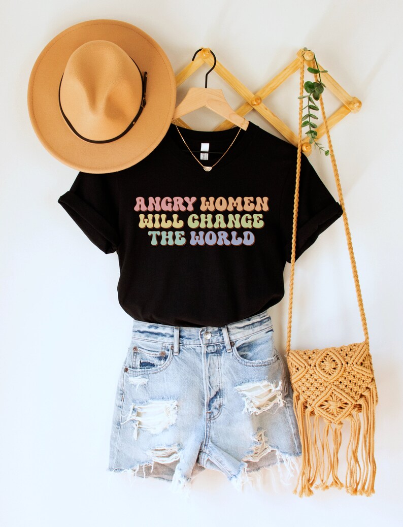 Może przedstawiać: Czarny T-shirt z napisem "Angry Women Will Change The World" w kolorach tęczy. Koszulka wisi na drewnianym wieszaku z brązowym kapeluszem i słomianą torbą.