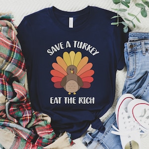 K&ouml;nnte beinhalten: Marineblaues T-Shirt mit einem Truthahn-Motiv und dem Text "SAVE A TURKEY EAT THE RICH". Der Truthahn hat einen bunten F&auml;cher aus Federn in Gelb-, Orange- und Rott&ouml;nen. Das Shirt wird mit blauen Jeans und wei&szlig;en Turnschuhen gezeigt.