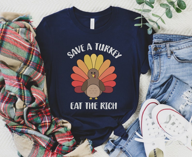 K&ouml;nnte beinhalten: Marineblaues T-Shirt mit einem Truthahn-Motiv und dem Text "SAVE A TURKEY EAT THE RICH". Der Truthahn hat einen bunten F&auml;cher aus Federn in Gelb-, Orange- und Rott&ouml;nen. Das Shirt wird mit blauen Jeans und wei&szlig;en Turnschuhen gezeigt.
