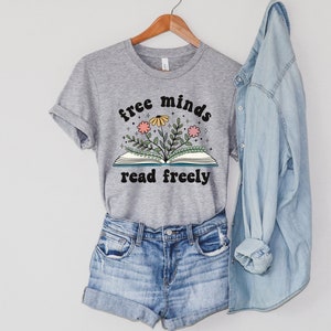 An athletic (medium grey) heater unisex crew neck short sleeve t-shirt with black text saying "free minds read freely" around a graphic of a book with colorful flowers and stars coming out of it