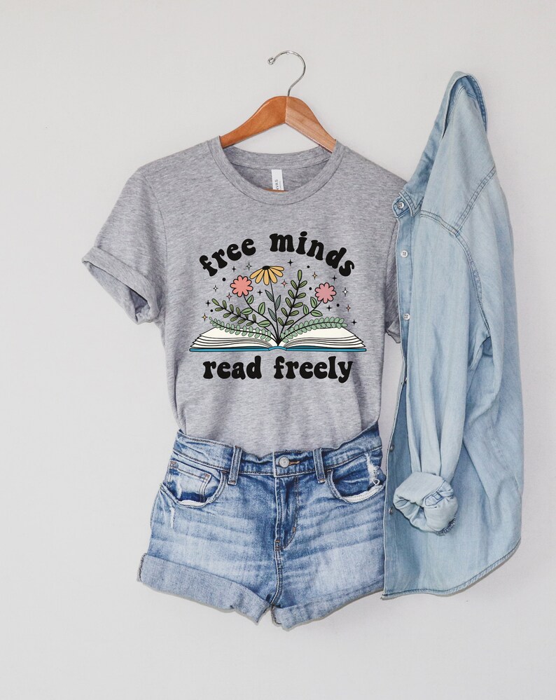 An athletic (medium grey) heater unisex crew neck short sleeve t-shirt with black text saying "free minds read freely" around a graphic of a book with colorful flowers and stars coming out of it