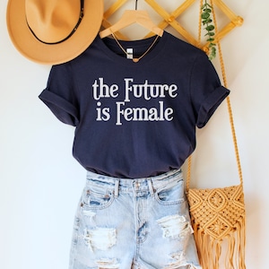 May include: A navy blue t-shirt with the text "the Future is Female" in white lettering. The shirt is hanging on a wooden hanger with a straw hat and a woven bag.