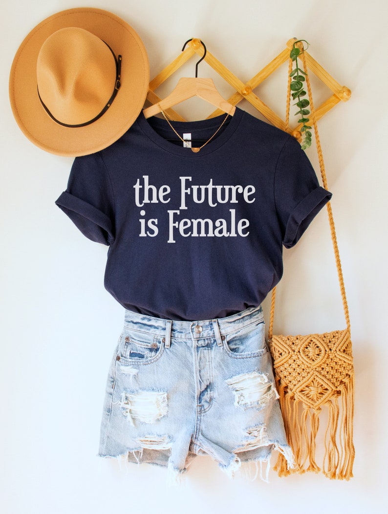 May include: A navy blue t-shirt with the text "the Future is Female" in white lettering. The shirt is hanging on a wooden hanger with a straw hat and a woven bag.