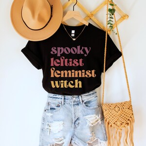 May include: Black t-shirt with the text "spooky leftist feminist witch" in a gradient of pink, orange, and yellow. The shirt is worn with denim shorts and a brown crocheted bag.