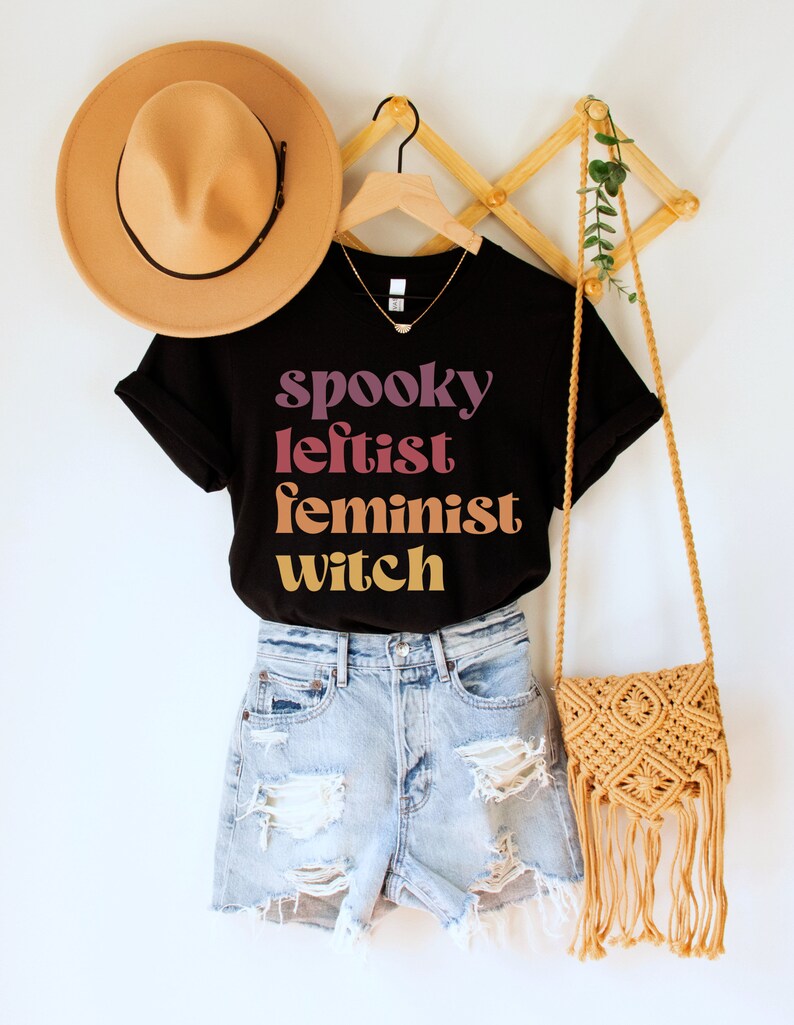 May include: Black t-shirt with the text "spooky leftist feminist witch" in a gradient of pink, orange, and yellow. The shirt is worn with denim shorts and a brown crocheted bag.