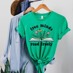 A heather kelly green unisex crew neck short sleeve t-shirt with black text saying "free minds read freely" around a graphic of a book with colorful flowers and stars coming out of it