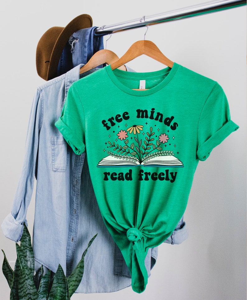 A heather kelly green unisex crew neck short sleeve t-shirt with black text saying "free minds read freely" around a graphic of a book with colorful flowers and stars coming out of it