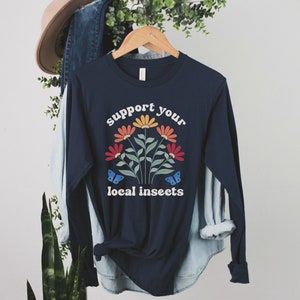 A navy blue unisex long sleeve crew neck t-shirt with the words "support your local insects" in white text around an image of colorful flowers and butterflies
