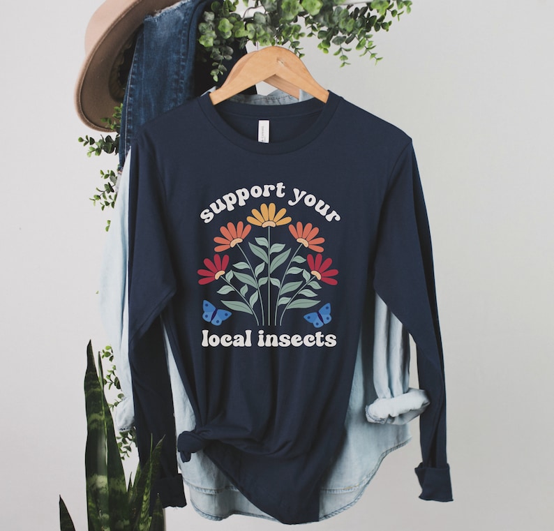 A navy blue unisex long sleeve crew neck t-shirt with the words "support your local insects" in white text around an image of colorful flowers and butterflies