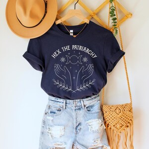 A navy blue unisex crew neck short sleeve Bella+Canvas t-shirt that says "hex the patriarchy" in a white uppercase distressed font, above a witchy white graphic with leaves, hands, stars, and a three moon symbol
