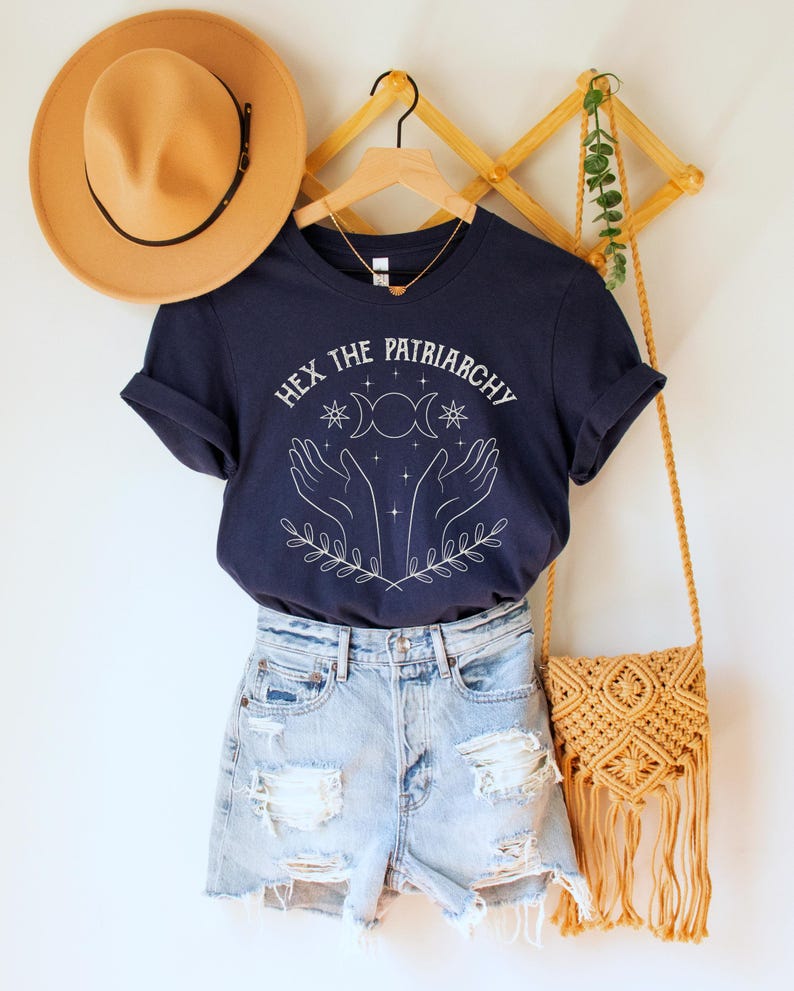 A navy blue unisex crew neck short sleeve Bella+Canvas t-shirt that says "hex the patriarchy" in a white uppercase distressed font, above a witchy white graphic with leaves, hands, stars, and a three moon symbol