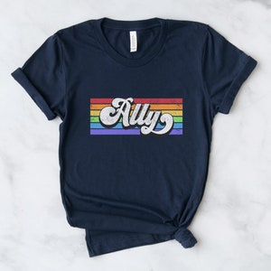 May include: Navy blue t-shirt with a rainbow stripe design and the word "Ally" in a retro font.