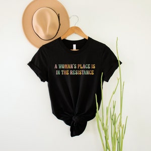 May include: Black t-shirt with the text "A woman's place is in the resistance" in rainbow colours.