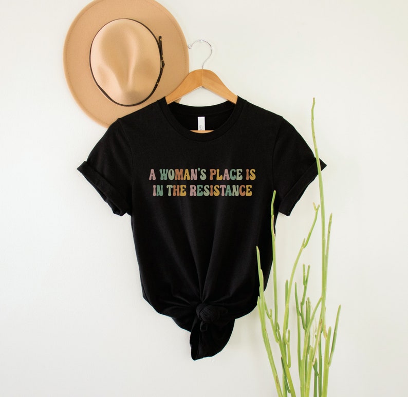 May include: Black t-shirt with the text "A woman's place is in the resistance" in rainbow colours.