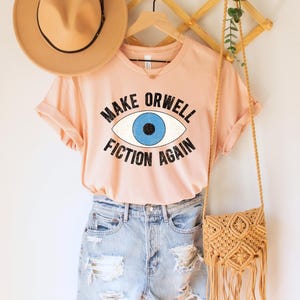 A heather peach unisex crew neck Bella Canvas brand t-shirt that says "MAKE ORWELL FICTION AGAIN" in black text, around an image of a blue eye. The graphic has a distressed vintaged look.