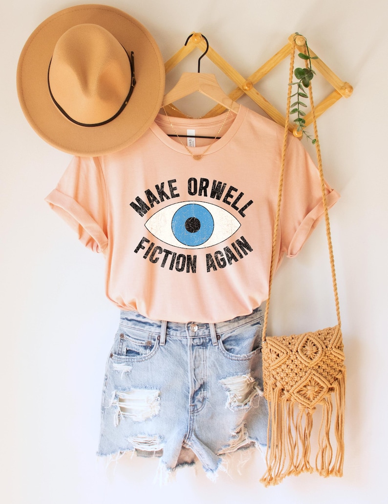 A heather peach unisex crew neck Bella Canvas brand t-shirt that says "MAKE ORWELL FICTION AGAIN" in black text, around an image of a blue eye. The graphic has a distressed vintaged look.