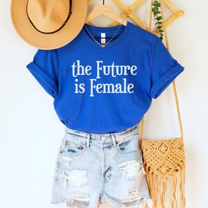May include: A royal blue t-shirt with the text "the Future is Female" in white. The t-shirt is hanging on a wooden hanger with a straw hat and a woven bag.