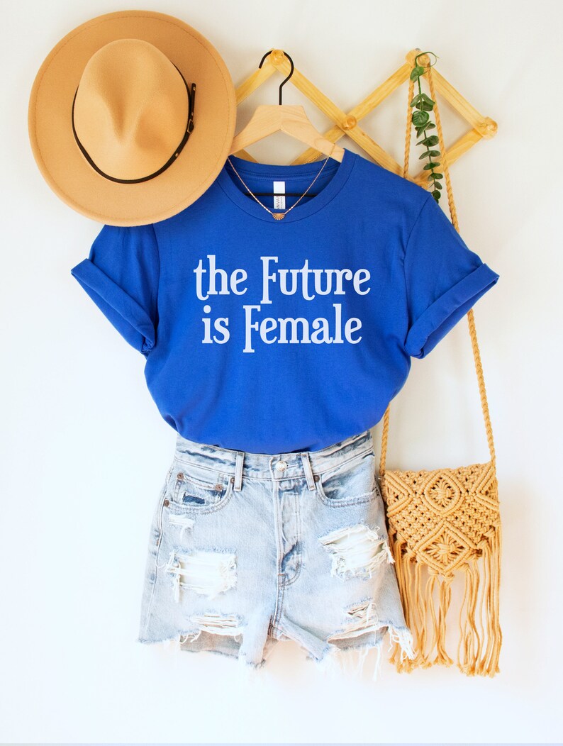 May include: A royal blue t-shirt with the text "the Future is Female" in white. The t-shirt is hanging on a wooden hanger with a straw hat and a woven bag.