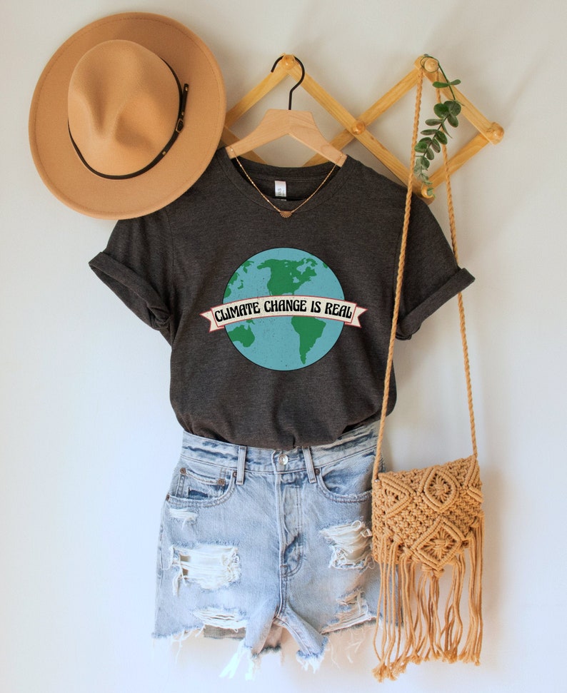 May include: A dark grey t-shirt with a graphic of the Earth and the text "Climate Change is Real" on a banner. The shirt is on a hanger with a brown hat and a woven bag.