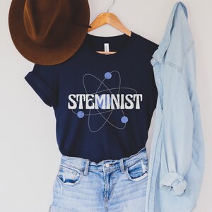 May include: A navy blue t-shirt with a white atom graphic and the text "STEMINIST" in white letters. The shirt is hanging on a wooden hanger with a brown hat and a light blue denim jacket.