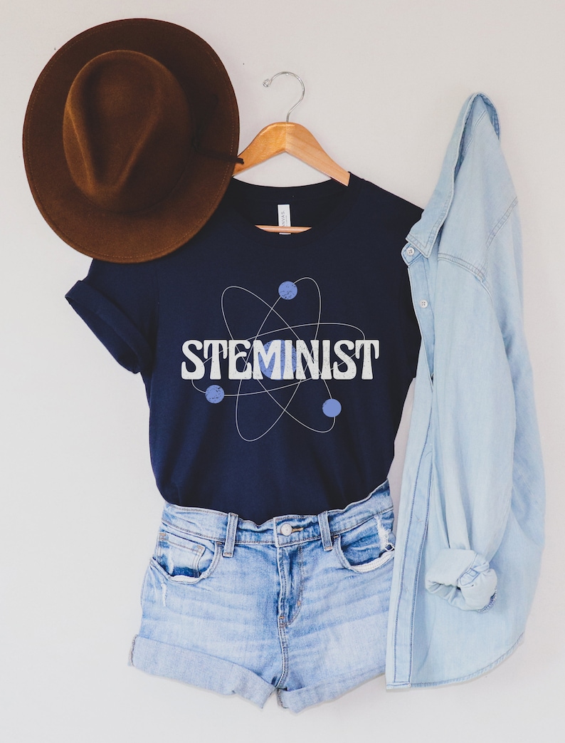 May include: A navy blue t-shirt with a white atom graphic and the text "STEMINIST" in white letters. The shirt is hanging on a wooden hanger with a brown hat and a light blue denim jacket.